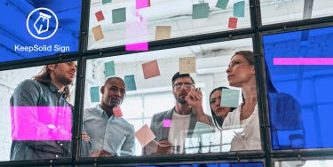 Group of young modern people in smart casual wear discussing IT sales solutions and using adhesive notes while standing behind the glass wall in the board room